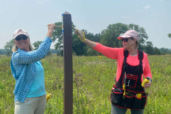 Two women paint yellow blazes on signage post in a green prairie.