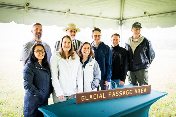 A photograph of seven people standing behind a sign reading "Glacial Passage Area" under a white tent.