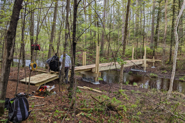 In the distance in a forest, two volunteers stand next to a nearly completed boardwalk that goes over a creek.