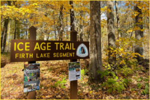 A brown Trailhead sign with yellow font reads: "Ice Age Trail Firth Lake Segment." An array of trees with fall colors stand in the backdrop.