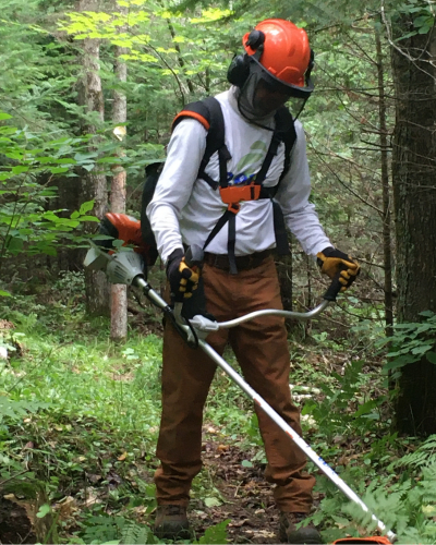 A volunteer in protective gear uses a brush cutter along a trail.