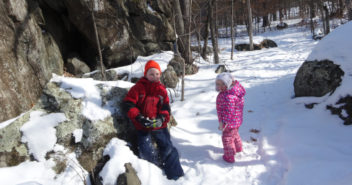 Two kids in snowsuits walk in the snow along a trail lined with boulders on a winter day.